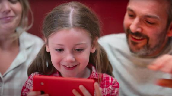 a grinning child peering into a mobile phone with her parents